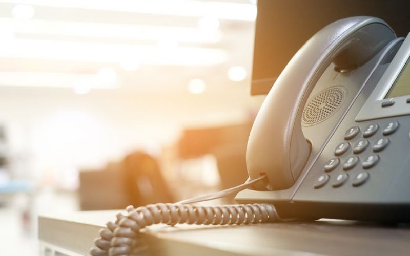 A VoIP phone sitting on the edge of the of a table in warm lighting.